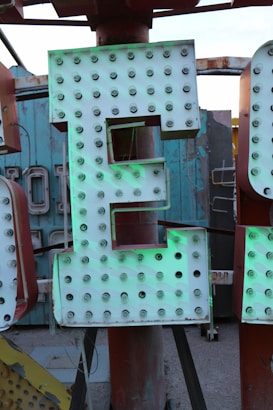 A large illuminated sign featuring a letter 'E', adorned with numerous bulbs, set against a rustic and slightly worn backdrop. The sign exhibits an industrial feel with hints of oxidation and weathering.