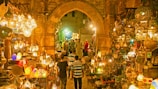 A vibrant marketplace scene featuring a variety of intricately designed lanterns displayed in an aisle with stone arches. The lighting creates a warm, golden glow throughout the space. Several people are seen browsing the market, with some walking along the cobblestone path surrounded by an array of colorful and ornate hanging lamps and decorations.