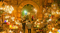 A vibrant marketplace scene featuring a variety of intricately designed lanterns displayed in an aisle with stone arches. The lighting creates a warm, golden glow throughout the space. Several people are seen browsing the market, with some walking along the cobblestone path surrounded by an array of colorful and ornate hanging lamps and decorations.