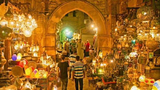 A vibrant marketplace scene featuring a variety of intricately designed lanterns displayed in an aisle with stone arches. The lighting creates a warm, golden glow throughout the space. Several people are seen browsing the market, with some walking along the cobblestone path surrounded by an array of colorful and ornate hanging lamps and decorations.
