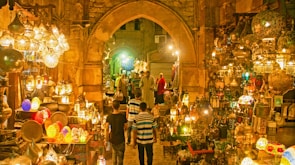 A vibrant marketplace scene featuring a variety of intricately designed lanterns displayed in an aisle with stone arches. The lighting creates a warm, golden glow throughout the space. Several people are seen browsing the market, with some walking along the cobblestone path surrounded by an array of colorful and ornate hanging lamps and decorations.