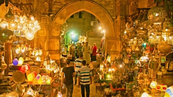A vibrant marketplace scene featuring a variety of intricately designed lanterns displayed in an aisle with stone arches. The lighting creates a warm, golden glow throughout the space. Several people are seen browsing the market, with some walking along the cobblestone path surrounded by an array of colorful and ornate hanging lamps and decorations.