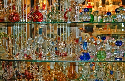 A collection of colorful glass and crystal items is displayed on multiple shelves. The objects vary in shape and include bottles, animal figurines, and decorative pieces. The glassware reflects light, creating a shimmering effect.