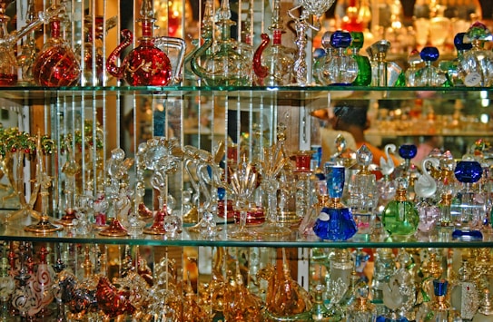 A collection of colorful glass and crystal items is displayed on multiple shelves. The objects vary in shape and include bottles, animal figurines, and decorative pieces. The glassware reflects light, creating a shimmering effect.