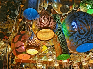 A vibrant array of intricately designed hanging lamps and lanterns in various shapes and sizes. The lamps are adorned with detailed perforations, allowing colorful light to filter through. The scene includes a mix of gold, blue, and green hues, creating an ornate and lively atmosphere with a craftsman or market vibe.