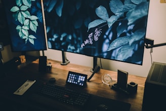 A workspace featuring two large computer monitors displaying images of green leaves and foliage. On the wooden desk, there's a keyboard, a small control pad, a notepad with a pen, and various electronic devices. The lighting is dim, contributing to a cozy and focused atmosphere.