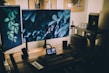 A cozy and stylish home office setup featuring a wooden desk with multiple monitors displaying nature-themed backgrounds. The desk is equipped with a keyboard, a note pad, a speaker, and other tech gadgets. There's soft, ambient lighting with potted plants and decorative items on a pegboard above the desk.
