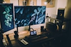 A cozy and stylish home office setup featuring a wooden desk with multiple monitors displaying nature-themed backgrounds. The desk is equipped with a keyboard, a note pad, a speaker, and other tech gadgets. There's soft, ambient lighting with potted plants and decorative items on a pegboard above the desk.