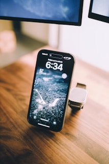 A smartphone with a nature-themed lock screen sits on a wooden surface. Nearby, a smartwatch rests on a stand. The setting appears to be a part of a workspace with a section of a monitor visible in the background.