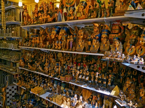 A collection of various Egyptian-themed statues and artifacts are arranged on several shelves. This includes busts of pharaohs, small pyramid models, and figures of ancient Egyptian gods and goddesses, all characterized by elaborate and colorful designs.
