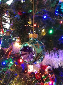 A detailed close-up of a decorated Christmas tree featuring a colorful glass ornament. The ornament is spherical with intricate designs, including gold accents and a prominent purple center. It hangs from a golden string amidst green pine needles. The background displays various colorful lights and tinsel, enhancing the festive atmosphere.