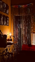 A warmly lit room with a vintage ambiance, featuring black and white photographs on the wall. A glowing lamp sits on a console table with potted plants, and a curtain of shimmering string lights hangs over a window or doorway.