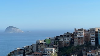 A sprawling cluster of favela-style buildings is perched on a hillside overlooking a vast, calm ocean. In the background, two rocky islands rise from the water under a clear blue sky. The buildings are densely packed and display a variety of colors, including natural tones, with some sections visibly weathered.