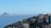 A sprawling cluster of favela-style buildings is perched on a hillside overlooking a vast, calm ocean. In the background, two rocky islands rise from the water under a clear blue sky. The buildings are densely packed and display a variety of colors, including natural tones, with some sections visibly weathered.