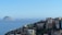 A sprawling cluster of favela-style buildings is perched on a hillside overlooking a vast, calm ocean. In the background, two rocky islands rise from the water under a clear blue sky. The buildings are densely packed and display a variety of colors, including natural tones, with some sections visibly weathered.