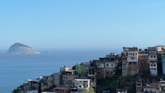 A sprawling cluster of favela-style buildings is perched on a hillside overlooking a vast, calm ocean. In the background, two rocky islands rise from the water under a clear blue sky. The buildings are densely packed and display a variety of colors, including natural tones, with some sections visibly weathered.