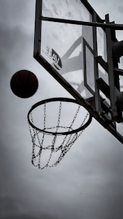 A basketball is captured in motion as it nears a hoop fitted with a metal chain net. The perspective is underneath the hoop, highlighting the angular structure of the backboard. The background features an overcast sky, creating a dramatic contrast with the dark silhouette of the basketball and hoop.
