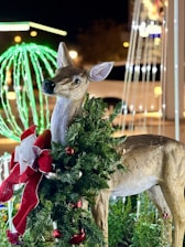 A lifelike deer sculpture is adorned with a festive green garland wrapped around its neck, decorated with red and silver ornaments. In the background, there are illuminated holiday decorations, including a green, glowing orb and other lights, creating a cheerful, seasonal atmosphere.