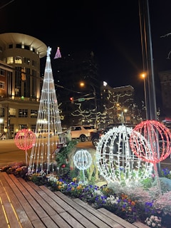 A nighttime street scene featuring festive holiday decorations. Illuminated structures in the shape of trees and spheres are adorned with white and red lights, surrounded by colorful flowers. In the background, a mix of modern and traditional buildings are highlighted with decorative lighting. A reindeer sculpture with holiday adornments is visible near the lights, and a few cars drive by on the street.