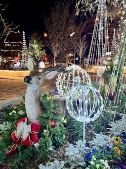 A festive outdoor scene featuring a decorative reindeer with illuminated antlers surrounded by colorful flowers and greenery. Bright festive lights, including wireframe spheres and tree decorations, illuminate the area at night. Visible in the background are buildings adorned with lights and leafless trees, all contributing to a holiday atmosphere.