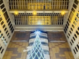 A large, illuminated Christmas tree stands indoors, surrounded by tall structures. The tree is brightly decorated with lights and is topped with a glowing star. Overhead, multiple yellow lights and windows are visible, and the surroundings include patterns and architectural details.