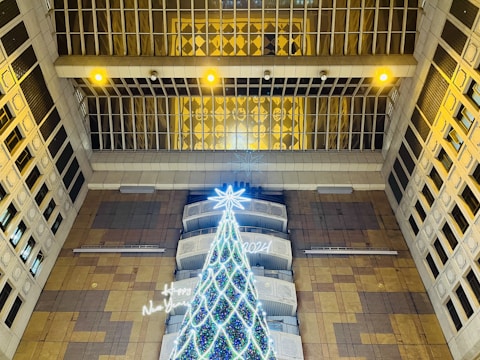 A large, illuminated Christmas tree stands indoors, surrounded by tall structures. The tree is brightly decorated with lights and is topped with a glowing star. Overhead, multiple yellow lights and windows are visible, and the surroundings include patterns and architectural details.