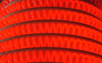 A close-up of bright orange and red coiled structures, possibly heating elements, arranged in parallel curves. The glowing loops create a vibrant and visually striking pattern against the darker background.