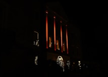 The image features a large building with prominent columns, illuminated by a warm orange-red light. Several windows are visible, glowing softly from interior lights. Decorative elements, likely holiday-themed, adorn the entrance with small trees or shrubs enhanced by white lights.