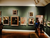 An art gallery displaying several framed paintings on a dark green wall. The paintings vary in size and depict people in different postures and attire. There are people observing the artworks and walking through the gallery. The lighting creates a warm and intimate atmosphere.