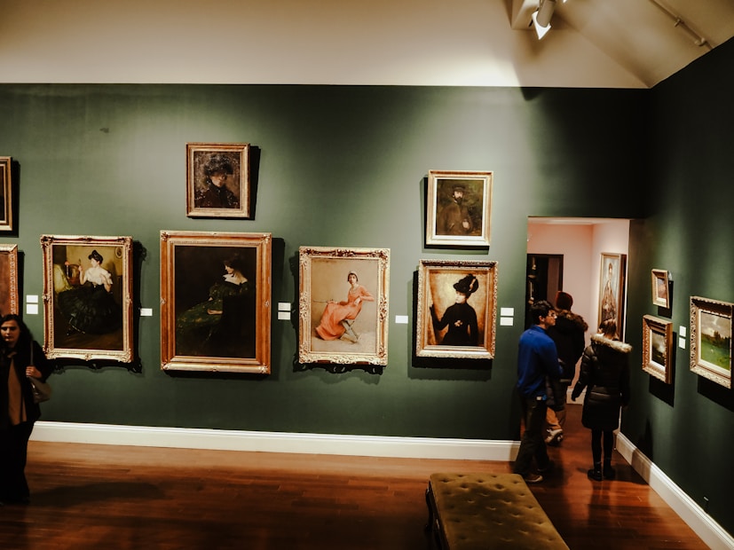 An art gallery displaying several framed paintings on a dark green wall. The paintings vary in size and depict people in different postures and attire. There are people observing the artworks and walking through the gallery. The lighting creates a warm and intimate atmosphere.