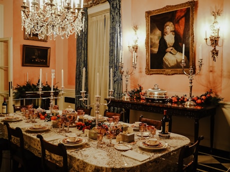 A luxurious dining room is set with an ornate table covered in a lace tablecloth and fine china. The table is adorned with candlesticks, glassware, and decorative fruits. A crystal chandelier hangs overhead, casting a warm glow. The walls are decorated with classical paintings and candelabras. Festive garlands with red berries and greenery add a touch of color to the sideboard.