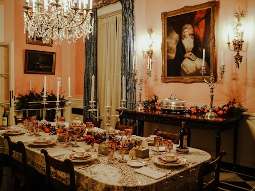 A luxurious dining room is set with an ornate table covered in a lace tablecloth and fine china. The table is adorned with candlesticks, glassware, and decorative fruits. A crystal chandelier hangs overhead, casting a warm glow. The walls are decorated with classical paintings and candelabras. Festive garlands with red berries and greenery add a touch of color to the sideboard.