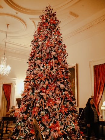 A large Christmas tree adorned with a variety of ornaments and red flowers stands prominently in a lavishly decorated room. The tree is beautifully lit and surrounded by upscale interior elements like chandeliers and plush curtains. A person is visible in the background near a lamp, adding a human element to the festive scene.