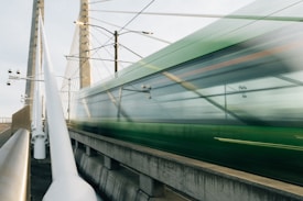 A green train speeds across a modern cable-stayed bridge, creating a motion blur effect. The bridge has tall, sturdy pillars with cables extending to support the structure. The scene captures a mix of urban infrastructure and movement.