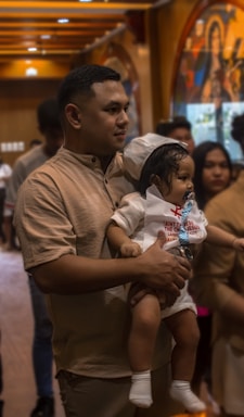 A man is holding a small child who is wearing a white outfit with red text on the front. The scene appears to be indoors with several people in the background and warm lighting from overhead. The child has a cap on and is in a secure grip by the man.