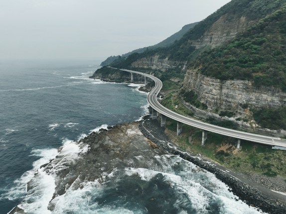 A scenic coastal road winds along a rocky cliffside with waves crashing against the shore. The lush vegetation blankets the towering cliffs, while the expansive ocean stretches into the horizon under an overcast sky.