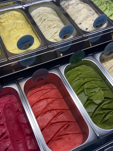 A display of various flavors of ice cream in metal containers, arranged in a freezer. Each container has a label indicating flavors like Durian, Honey Ginger, Vanilla Brownie, Strawberry, and Basil Lemon. The ice cream colors range from yellow, cream, green, to bright red and pink.