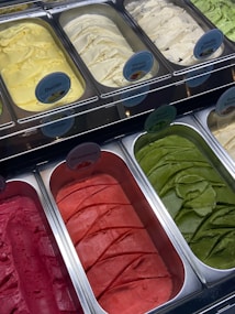 A display of various flavors of ice cream in metal containers, arranged in a freezer. Each container has a label indicating flavors like Durian, Honey Ginger, Vanilla Brownie, Strawberry, and Basil Lemon. The ice cream colors range from yellow, cream, green, to bright red and pink.