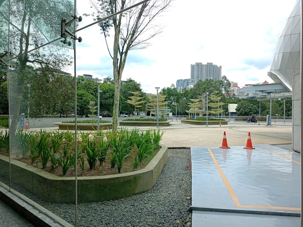 A modern urban environment with a glass façade reflecting nearby trees. In the foreground, there is a landscaped area with lush green plants bordered by pebbles. The paved path leads towards orange traffic cones indicating a restricted area. In the background, tall buildings and a few parked vehicles are visible under a cloudy sky.