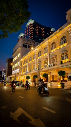 A bustling urban street scene at night with several people riding motorcycles on a road in front of an elegantly lit hotel. The hotel fa&ccedil;ade is illuminated with warm lights, showcasing its classical architecture. In the background, a modern high-rise building with red accent lights contrasts against the deep blue evening sky.