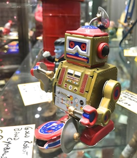 A vintage-style toy robot is displayed in a glass cabinet. The robot features a colorful design with a mix of metallic gold and vibrant red hues. It has an old-fashioned control panel with various dials and buttons on its chest, and it holds silver cymbals in its hands. The robot's eyes are noticeable with red and blue circular patterns, and it has an antenna on top of its head.