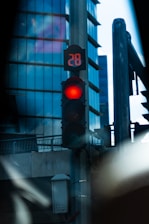 A traffic light displaying a red signal and a numeric countdown of 28, positioned against a backdrop of modern glass buildings. The scene is urban and features elements such as street lamps and metal structures.