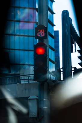 A traffic light displaying a red signal and a numeric countdown of 28, positioned against a backdrop of modern glass buildings. The scene is urban and features elements such as street lamps and metal structures.