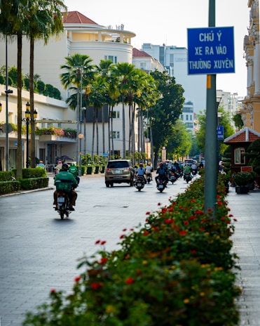 A bustling urban street lined with tall palm trees and neatly manicured hedges. Several motorbikes and a car are driving down the road, with a blue street sign in Vietnamese visible in the foreground. The architecture includes modern buildings with white facades and a few plants on the balconies. Bright sunlight casts long shadows, indicating a clear day.