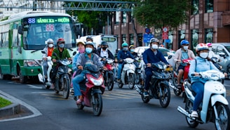 A group of people, wearing helmets and masks, ride scooters alongside a green bus on a city street. The scene is busy with traffic, and buildings can be seen in the background.