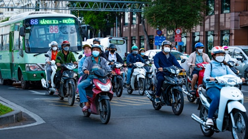 A group of people, wearing helmets and masks, ride scooters alongside a green bus on a city street. The scene is busy with traffic, and buildings can be seen in the background.