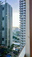 Two high-rise apartment buildings frame a view of a distant cityscape with skyscrapers, as sunset creates a bright light on the horizon. Below, a pool and palm trees are visible, suggesting an urban residential area with recreational facilities.