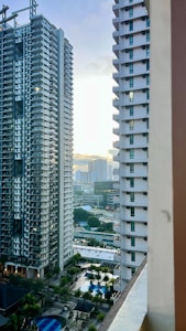 Two high-rise apartment buildings frame a view of a distant cityscape with skyscrapers, as sunset creates a bright light on the horizon. Below, a pool and palm trees are visible, suggesting an urban residential area with recreational facilities.