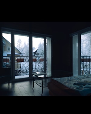 A room with large glass doors showcasing a snowy outdoor scene. A snowy landscape with trees and rooftops is visible outside. Inside, there is a glass table and part of a bed with a blanket. The floor is dark, possibly wooden or tiled.