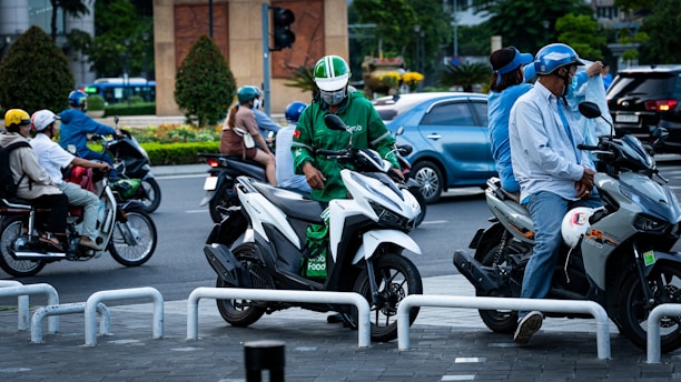 A busy urban street scene with multiple motorcyclists stopped at an intersection. The riders are wearing helmets, and one person is wearing a green jacket with a food delivery service logo on it. The environment includes cars and lush green trees in the background.